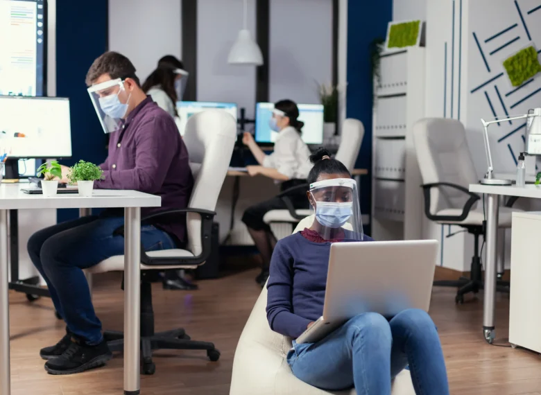 black-freelancer-with-protection-mask-against-coronavirus-sitting-armchair-middle-office-room-analysing-project-digital-tablet-multiethnic-business-team-working-respecting-social-distance